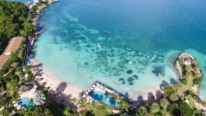 An aerial view of a resort with a clear blue sea, a sandy beach, and a swimming pool surrounded by palm trees. - Blue Waters Resort - Rock Cottage (Antigua and Barbuda)