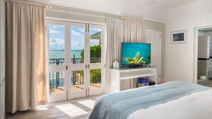 A hotel room with a large bed, a television on a stand, a sliding glass door leading to a balcony with a view of the sea, and a small bathroom with a shower. - Blue Waters Resort - Rock Cottage (Antigua and Barbuda)