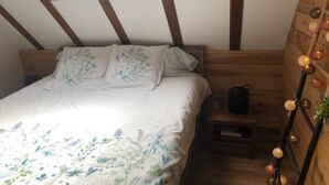 Un dormitorio con tragaluz, vigas de madera, una cama con ropa de cama floral, una mesita de noche y una lámpara fijada a la pared.