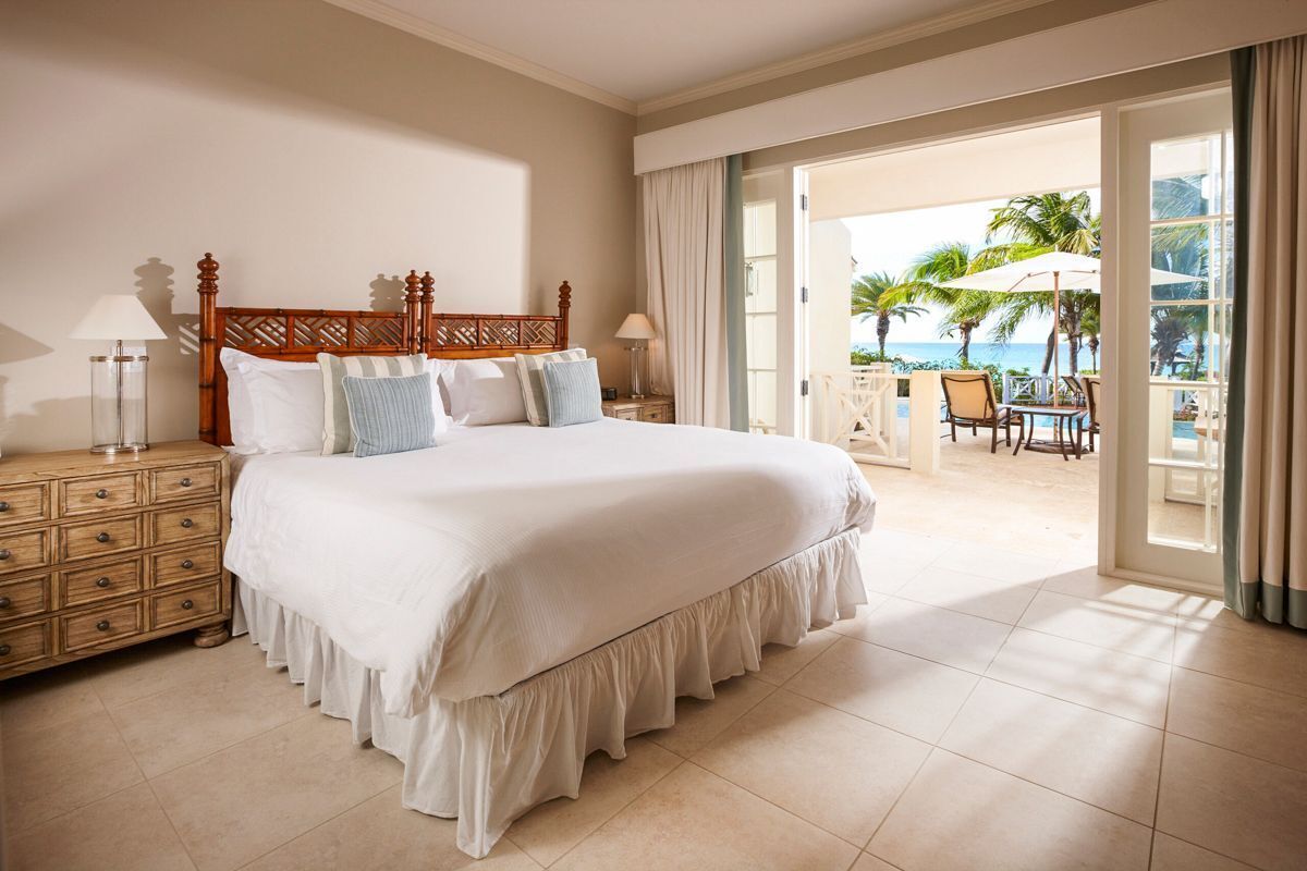 Une chambre d’hôtel avec un grand lit, une table de chevet en bois et une vue sur la plage par une porte ouverte.