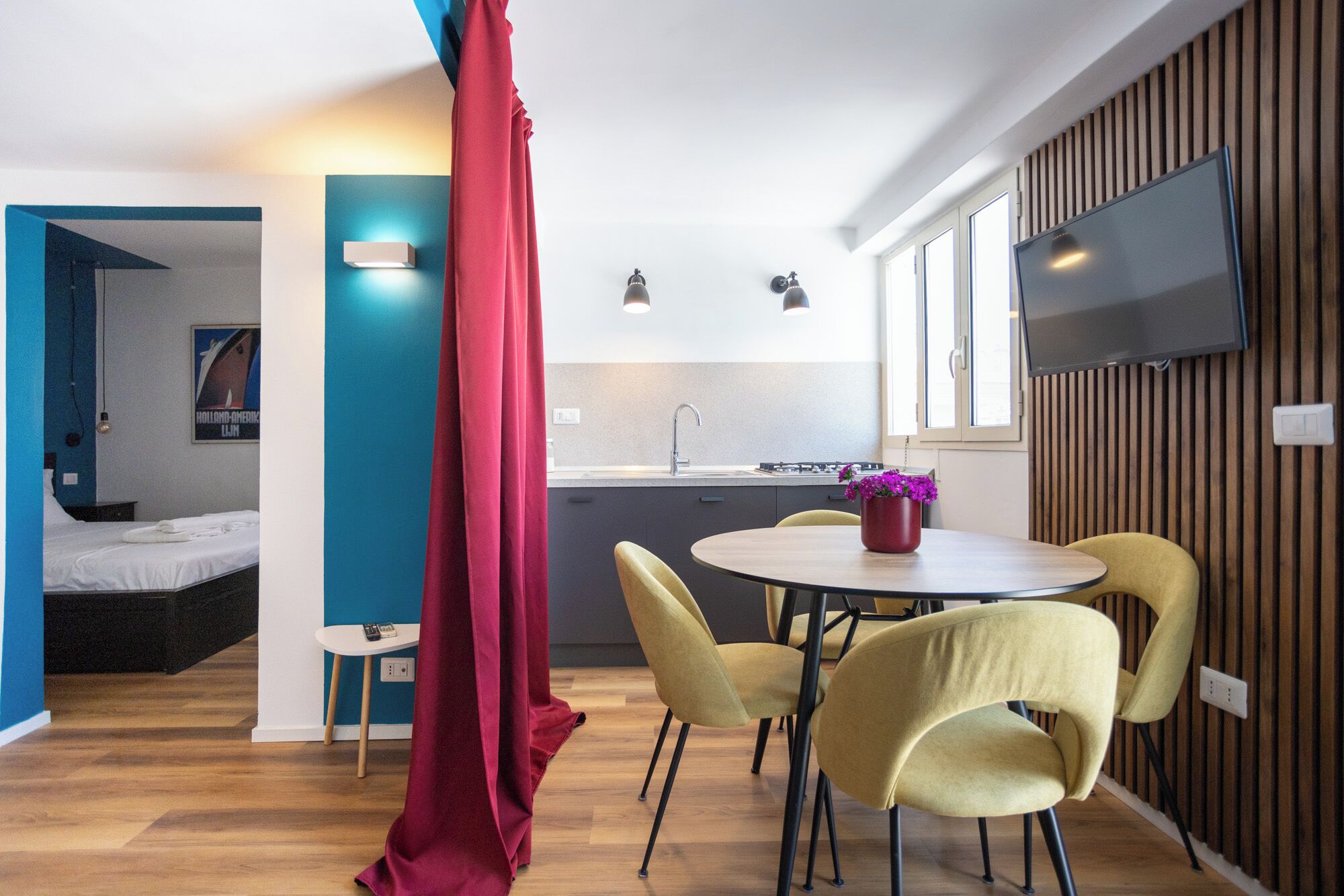 Una camera moderna con un letto, un'area pranzo con sedie, una cucina con lavandino e un televisore fissato al muro.