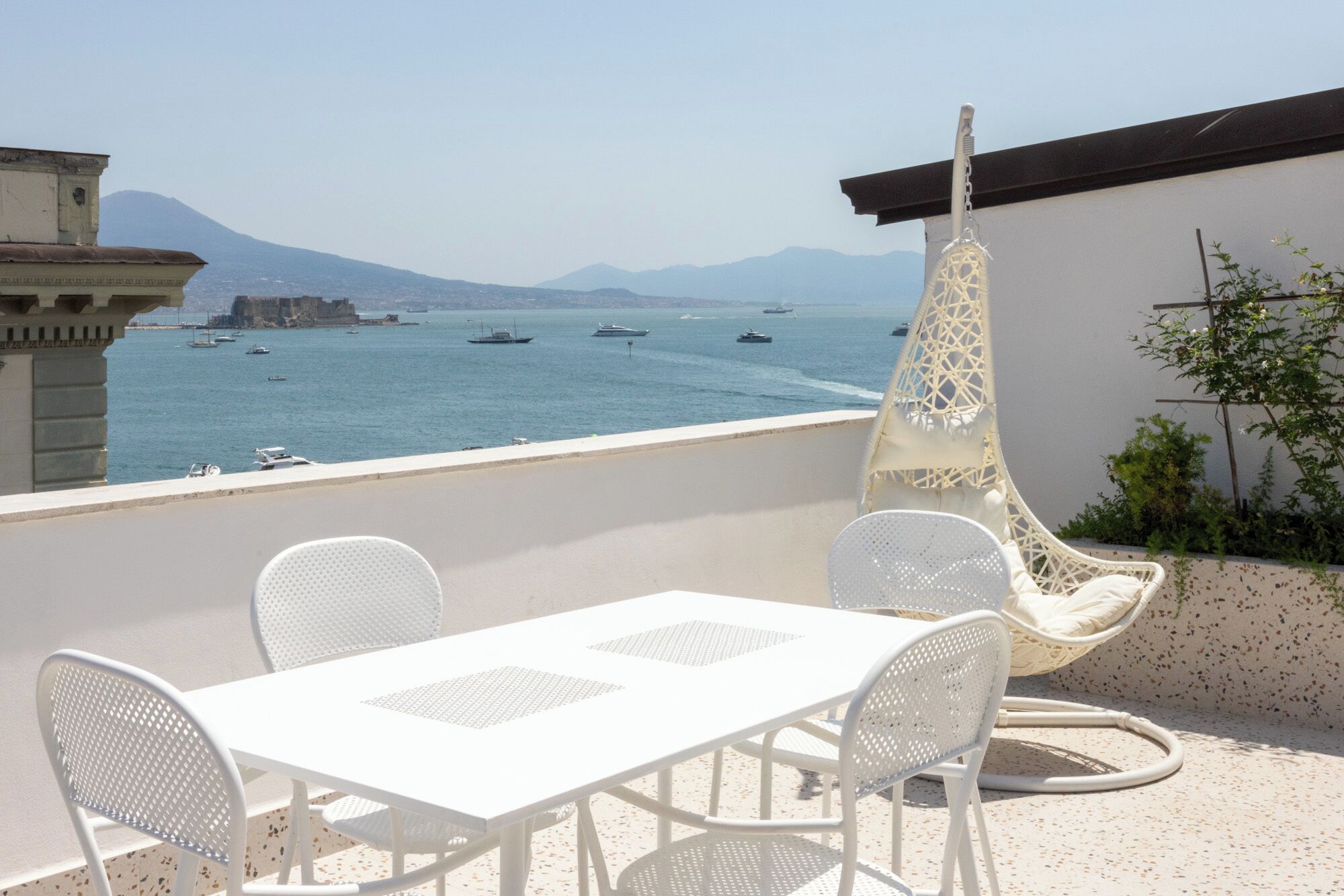 Una terrazza con tavolo e sedie bianche, una sedia a dondolo e vista sul mare con barche.