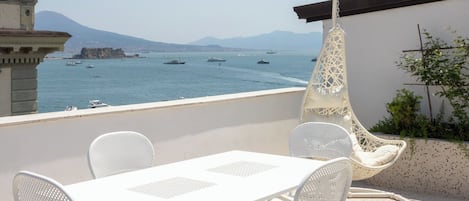 Une terrasse avec une table et des chaises blanches, un fauteuil suspendu et une vue sur la mer, où l’on aperçoit des bateaux.