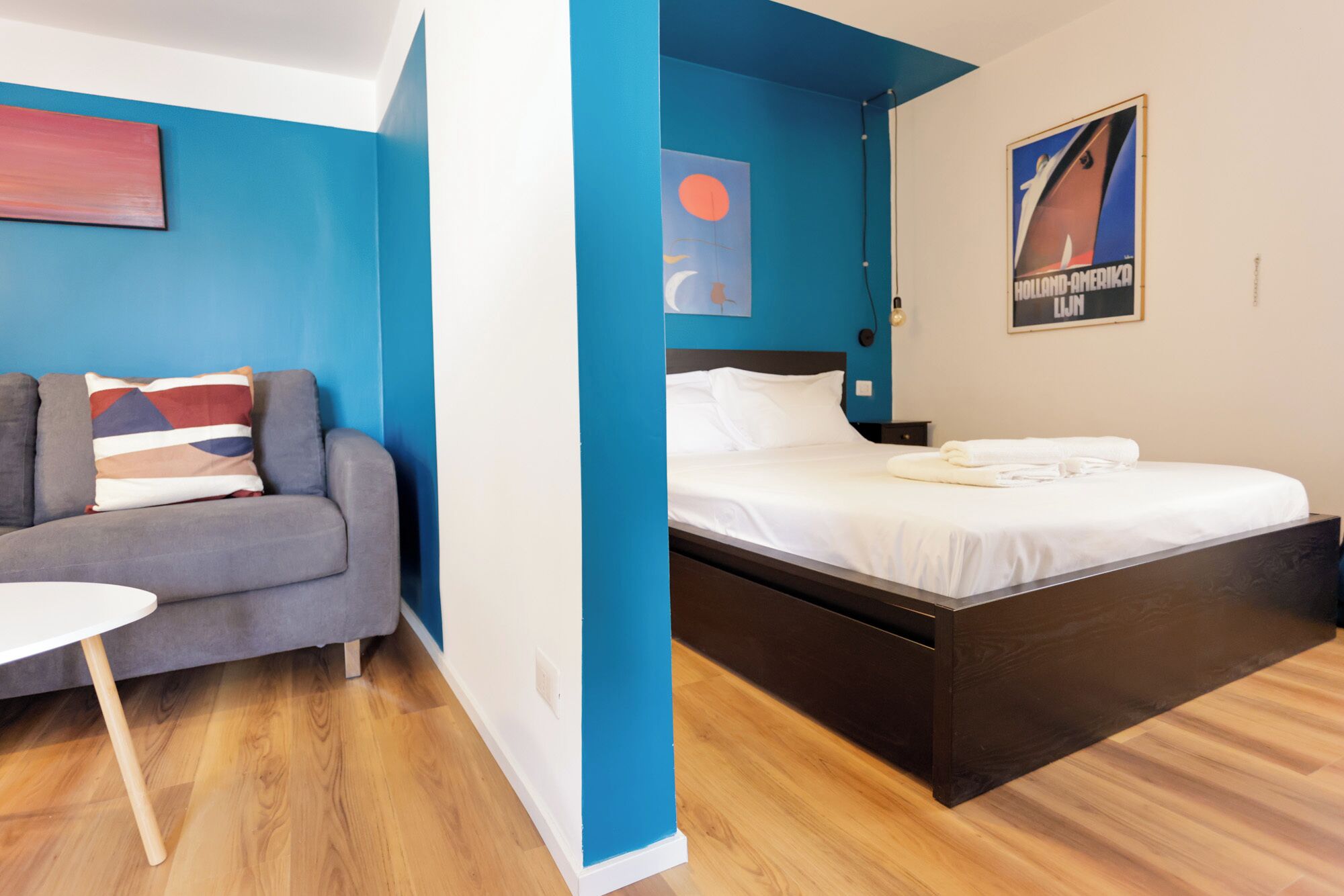 Una camera d'albergo con un letto, un divano grigio, un piccolo tavolino bianco e una parete azzurra.
