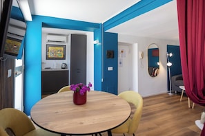 A room with a round wooden table, yellow chairs, a red vase with purple flowers, a kitchen area with a fridge and an air conditioner, and a mirror on the wall.