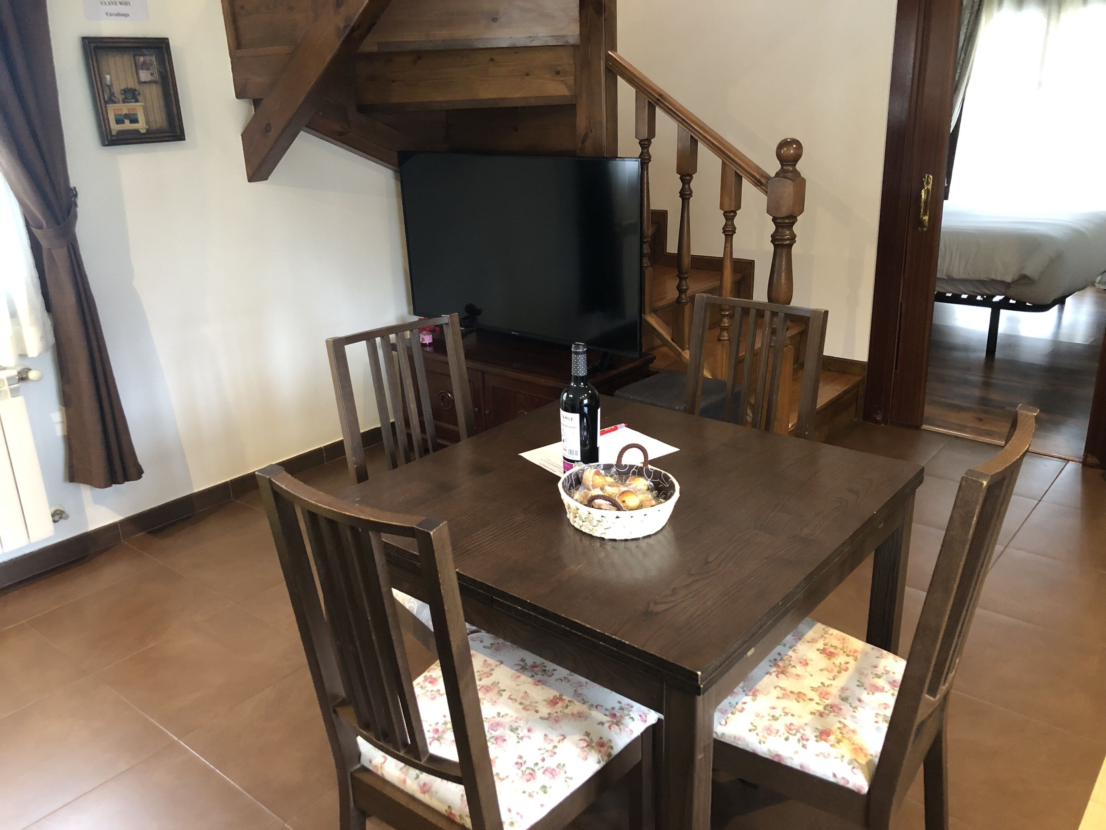 Un comedor con una mesa y sillas de madera, una cesta de pan, una botella de vino y un televisor al fondo.