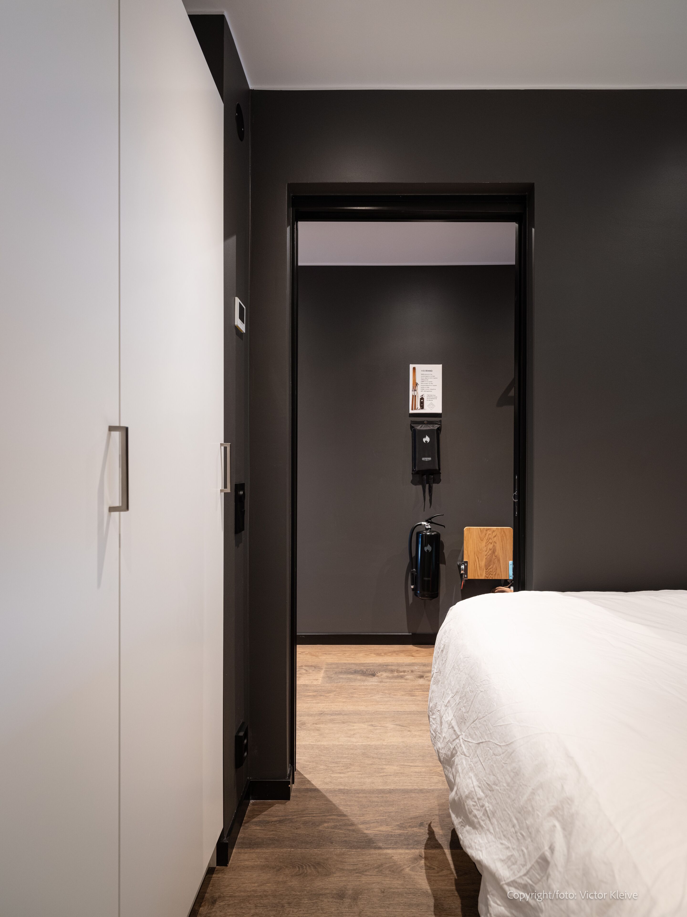 Hotellihuone, jossa on sänky, puulattia, ovi ja seinään kiinnitetty puhelin.