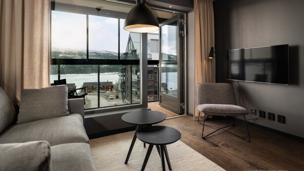 A modern living room with a large window, a sofa, a chair, a small table, and a TV mounted on the wall.