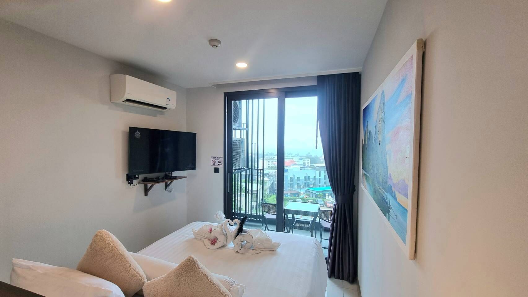 A hotel room with a bed, a TV, an air conditioner, a balcony with a table and chairs, and a painting on the wall.