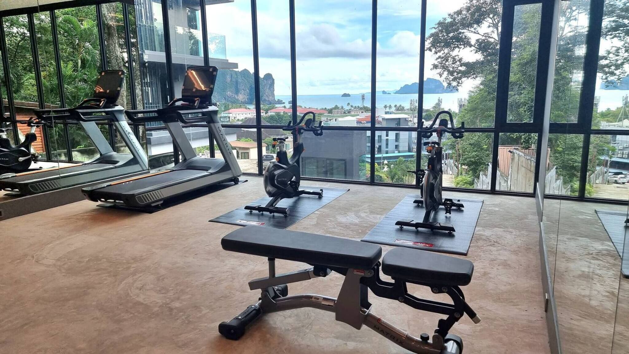 A gym with treadmills, an elliptical, and a bench press area, with large windows overlooking a cityscape.