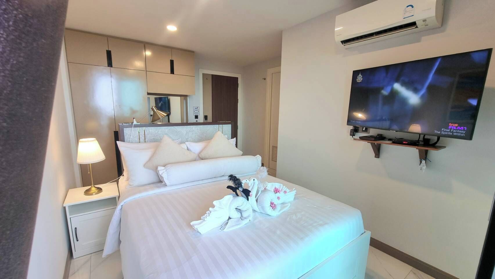 A hotel room with a bed, a TV mounted on the wall, a bedside lamp, and a wall-mounted air conditioner.
