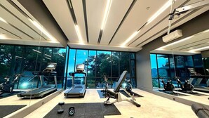Academia moderna com esteiras, bicicleta ergométrica e equipamentos de musculação. Grandes janelas proporcionam vista para árvores e céu claro.