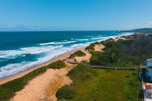 Un paisaje costero con una playa arenosa, un paseo de madera y abundante vegetación.