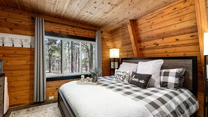 A cozy cabin bedroom with a wooden ceiling, a large window with curtains, a bed with checkered bedding, and a bedside table with a lamp.