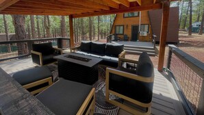 A covered outdoor deck with wooden furniture, a fire pit, and a view of a cabin in a wooded area.