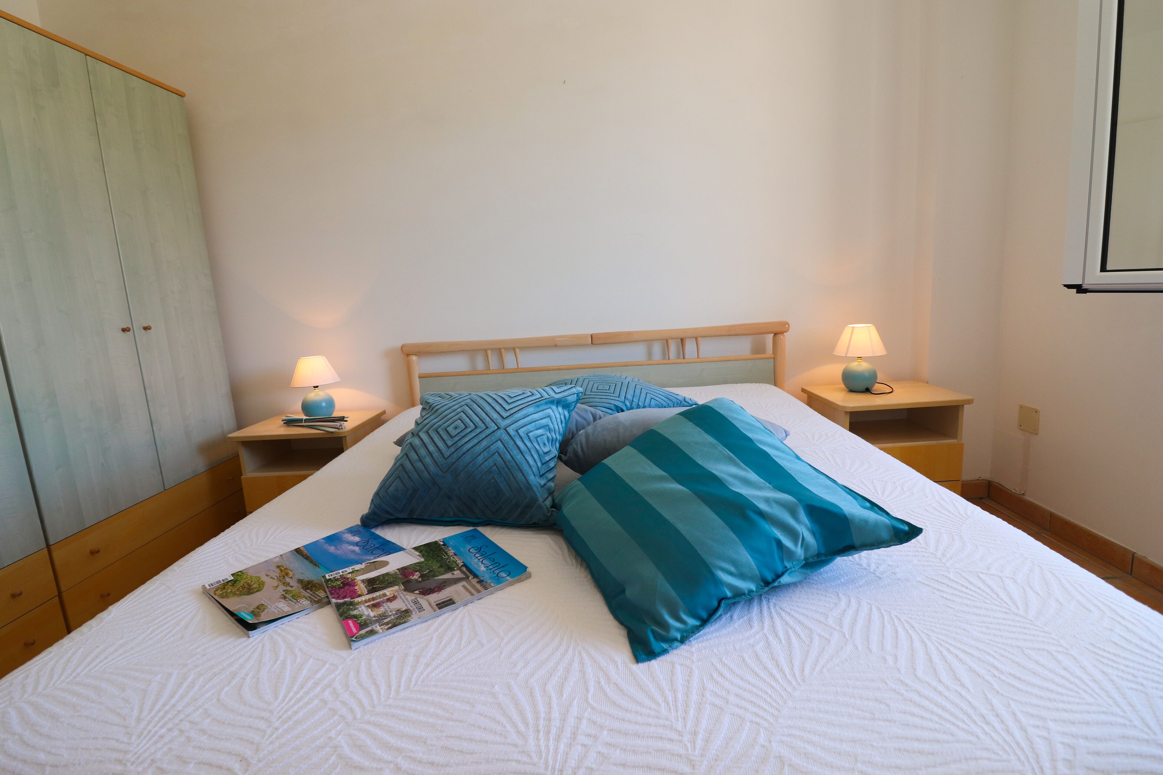 Un letto rifatto con cuscini blu e bianchi, due abat-jour e un comodino in legno con un libro e una penna.