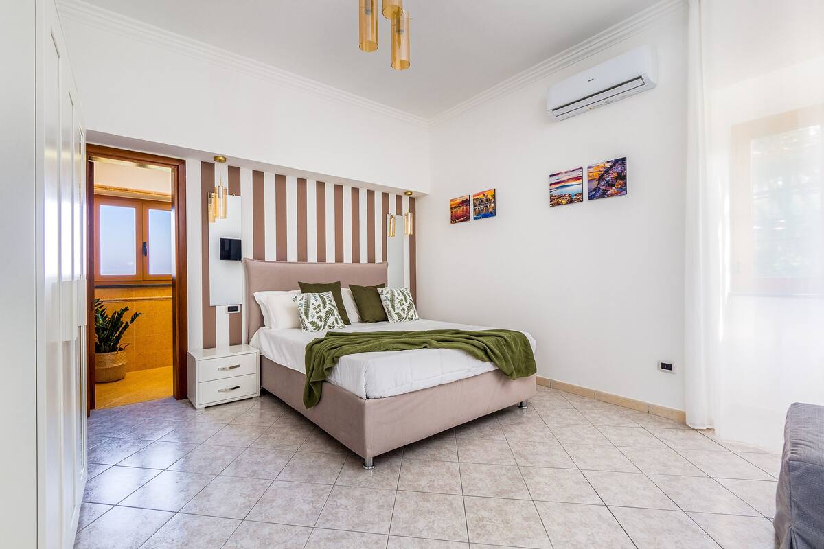 A modern bedroom with a bed, bedside tables, a wardrobe, a hanging light fixture, an air conditioning unit, and a window.