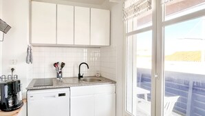 A modern kitchen with white cabinets, a granite countertop, a built-in dishwasher, and a window with a view of a balcony. - Spacious apartment with ocean view in the Chaume district (Les Sables-d´Olonne)