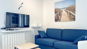 A modern living room with a blue sofa, a flat-screen TV mounted on a white shelf, a small table, and a painting of a beach walkway. - Spacious apartment with ocean view in the Chaume district (Les Sables-d´Olonne)