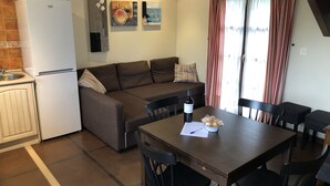 A compact kitchen with a refrigerator, a dining table with chairs, a sofa, and a window with striped curtains.