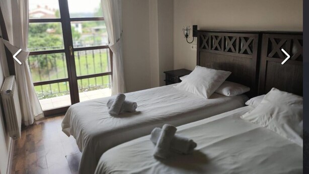 A hotel room with two beds, a view through a large window, and a nightstand with a lamp.