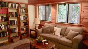 Ein gemütliches Wohnzimmer mit einem beigen Sofa, einem Holztisch mit Büchern und einer Pflanze, einem Bücherregal voller Bücher und einem Fenster mit Blick auf Bäume.