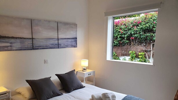 Una cama bien hecha con sábanas blancas, dos almohadas oscuras y una manta azul. Hay una mesita de noche con una lámpara, y una ventana con vistas a follaje y flores.