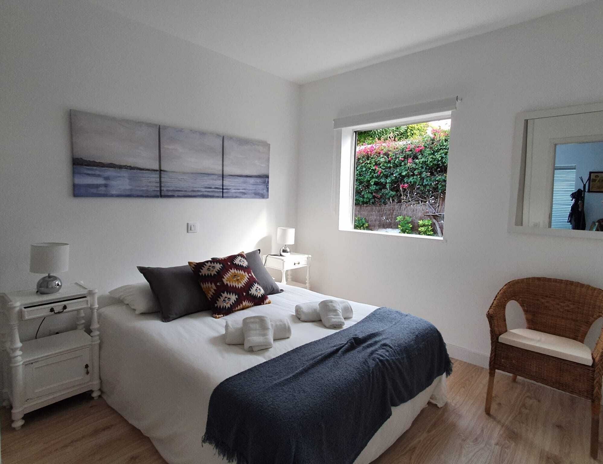 Un dormitorio con cama, una mesita de noche, una silla, una ventana que da a zonas verdes y un cuadro en la pared.