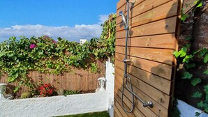 Puerta de madera con cabezal de ducha y manguera, pared blanca y jardín con plantas y flores verdes.