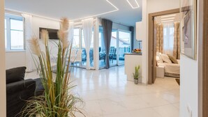 Una sala de estar moderna con un sofá negro, una mesa de centro y un comedor con sillas. Hay una zona de cocina con mostrador y un refrigerador. La habitación tiene ventanas amplias con cortinas e iluminación empotrada en el techo.