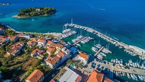 Vista aérea de un pueblo costero con un puerto deportivo, casas con techos rojos y una pequeña isla con vegetación.