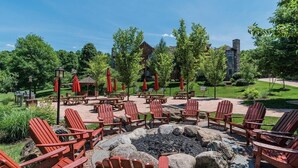 An outdoor seating area with Adirondack chairs and red umbrellas, a fire pit, and a view of a building with a stone chimney. - Little Getaway at Mountain Creek Sanctuary (Vernon Township)