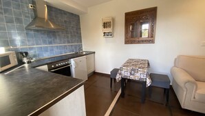 Eine kompakte Küche mit Mikrowelle, Kaffeemaschine, Herd und Geschirrspüler. Ein kleiner Tisch mit gemustertem Tischläufer und ein beiges Sofa sind ebenfalls vorhanden.