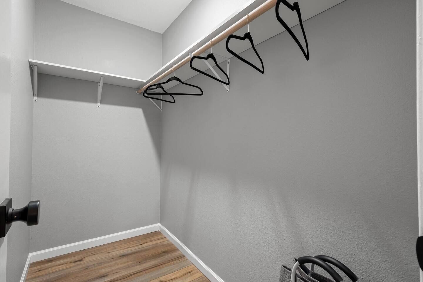 Un coin de pièce avec un plancher en bois, un mur blanc et un portant métallique avec des cintres noirs.