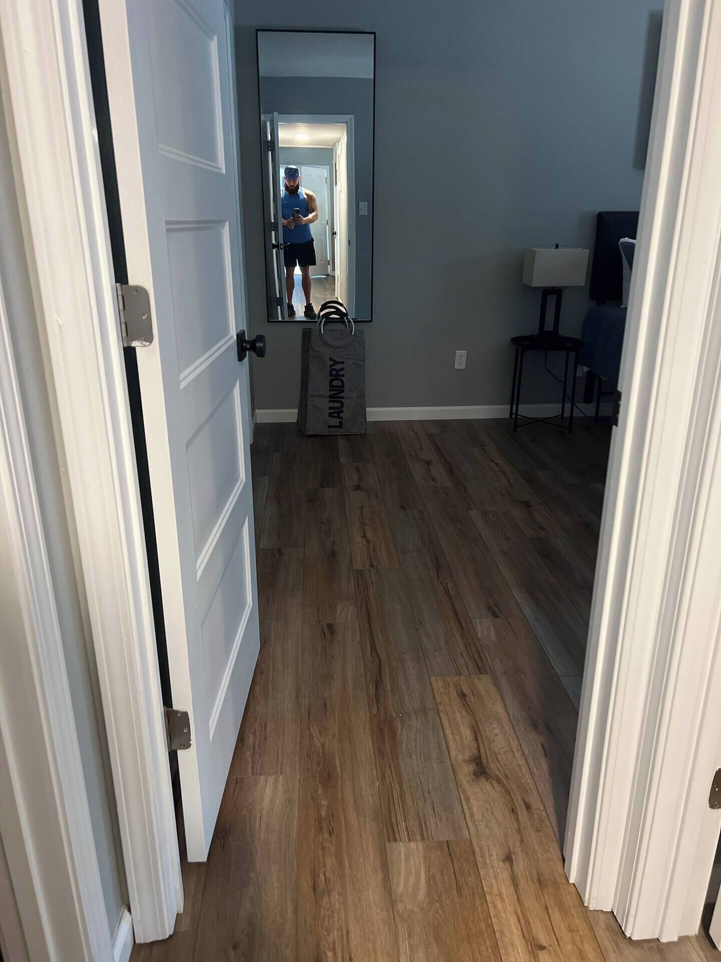 A hallway with wooden flooring, a mirror reflecting a person, and a grey 'laundry' bag on the floor.