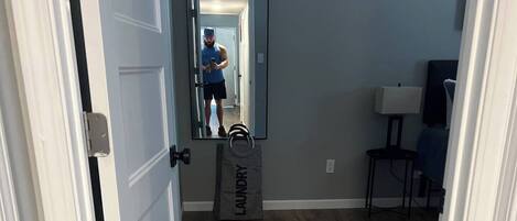 A hallway with wooden flooring, a mirror reflecting a person, and a grey 'laundry' bag on the floor.