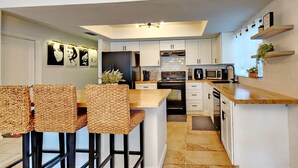 Una cocina moderna con armarios blancos, encimera de madera y horno empotrado. Hay dos taburetes de bar con asientos de mimbre y una planta pequeña en la encimera.