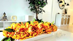 Una habitación con un sofá blanco, una pequeña estatua de Buda, una planta en maceta y un arreglo floral artificial grande sobre una mesa blanca.