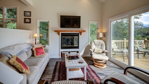 A cozy living room with a fireplace, comfortable seating, and large windows offering a view of the outdoors.