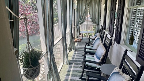 Een overdekte veranda met gestreepte vloer, zwarte houten stoelen met witte kussens en een hangplant.