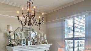 Een kamer met een witte open haard, een kroonluchter, een tafel met lampen en een raam met jaloezieën.