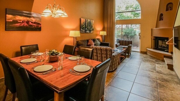 A dining area with a wooden table set for four, flanked by leather chairs. A chandelier hangs above, and a painting adorns the wall. A sofa and a fireplace are visible in the background. - Capital Vacations Sedona Springs Resort - 2 Bedroom (3-Night) (Sedona)