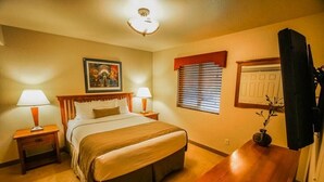 A hotel room with a large bed, two bedside tables with lamps, a wooden desk, a television, and a window with blinds.