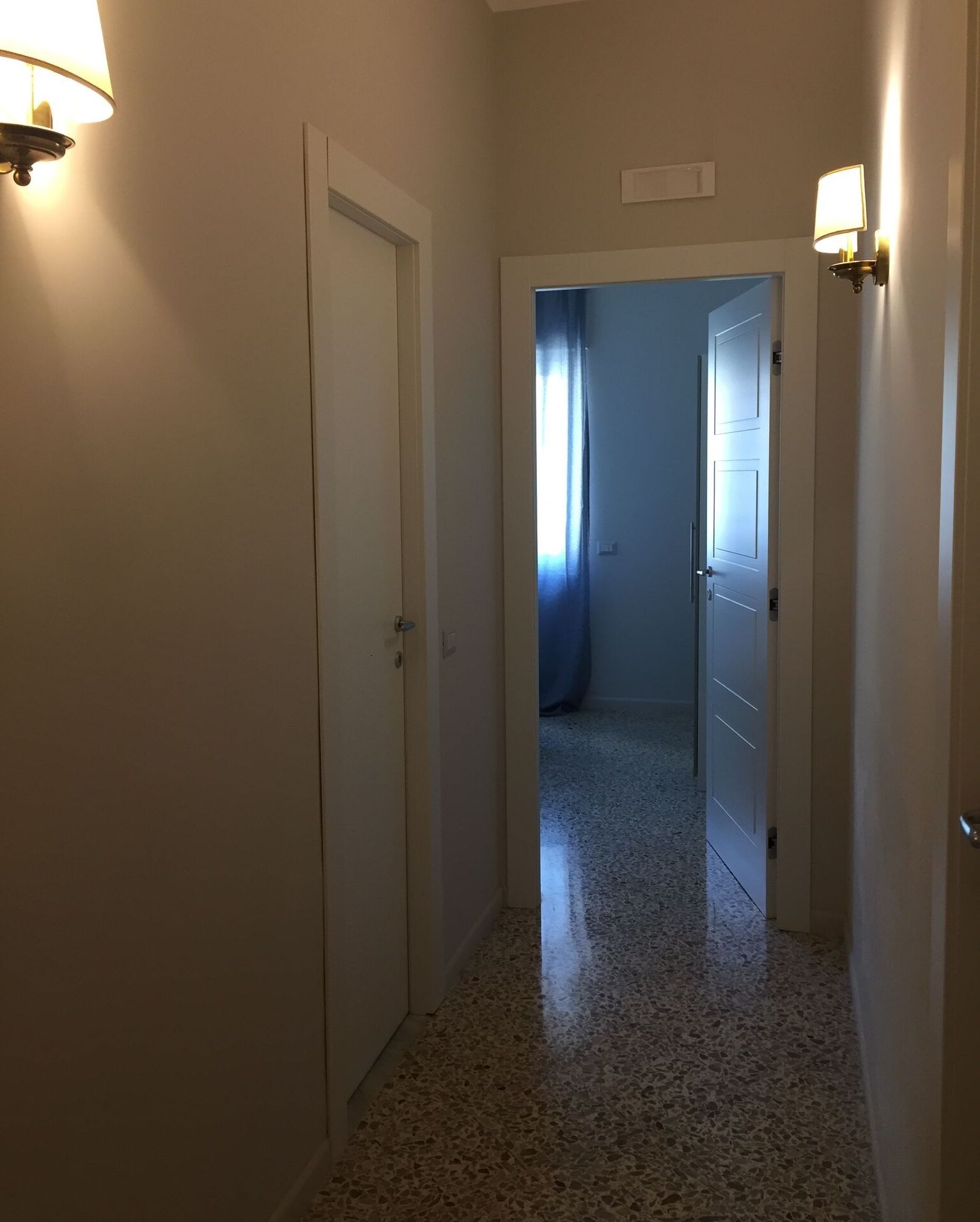 A narrow hallway with white walls and a speckled floor leading to a room with a door and a window with curtains.