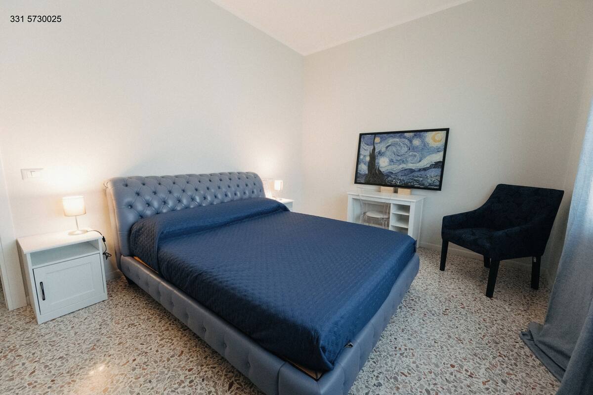 Un dormitorio con cama de cabecera capitonada, mesita de noche con lámpara, una silla y un cuadro en la pared.