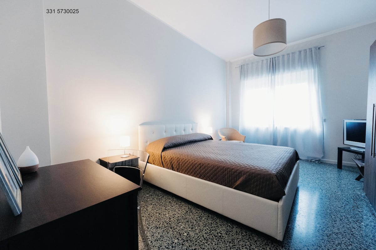 Una habitación de hotel moderna con cama, mesita de noche, lámpara, televisión y escritorio con silla.