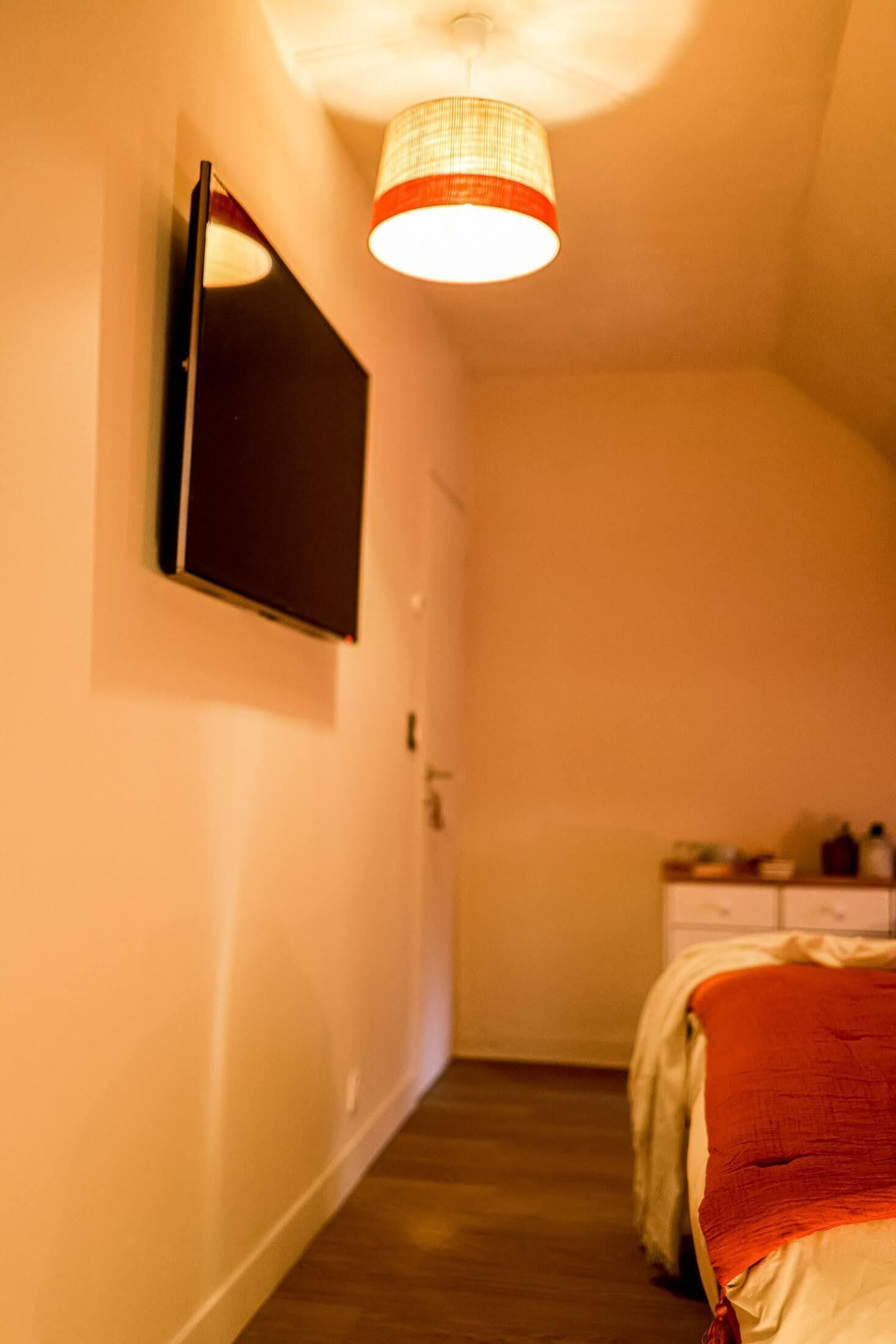 Um corredor estreito com uma cama à direita, um espelho à esquerda e um candeeiro suspenso.