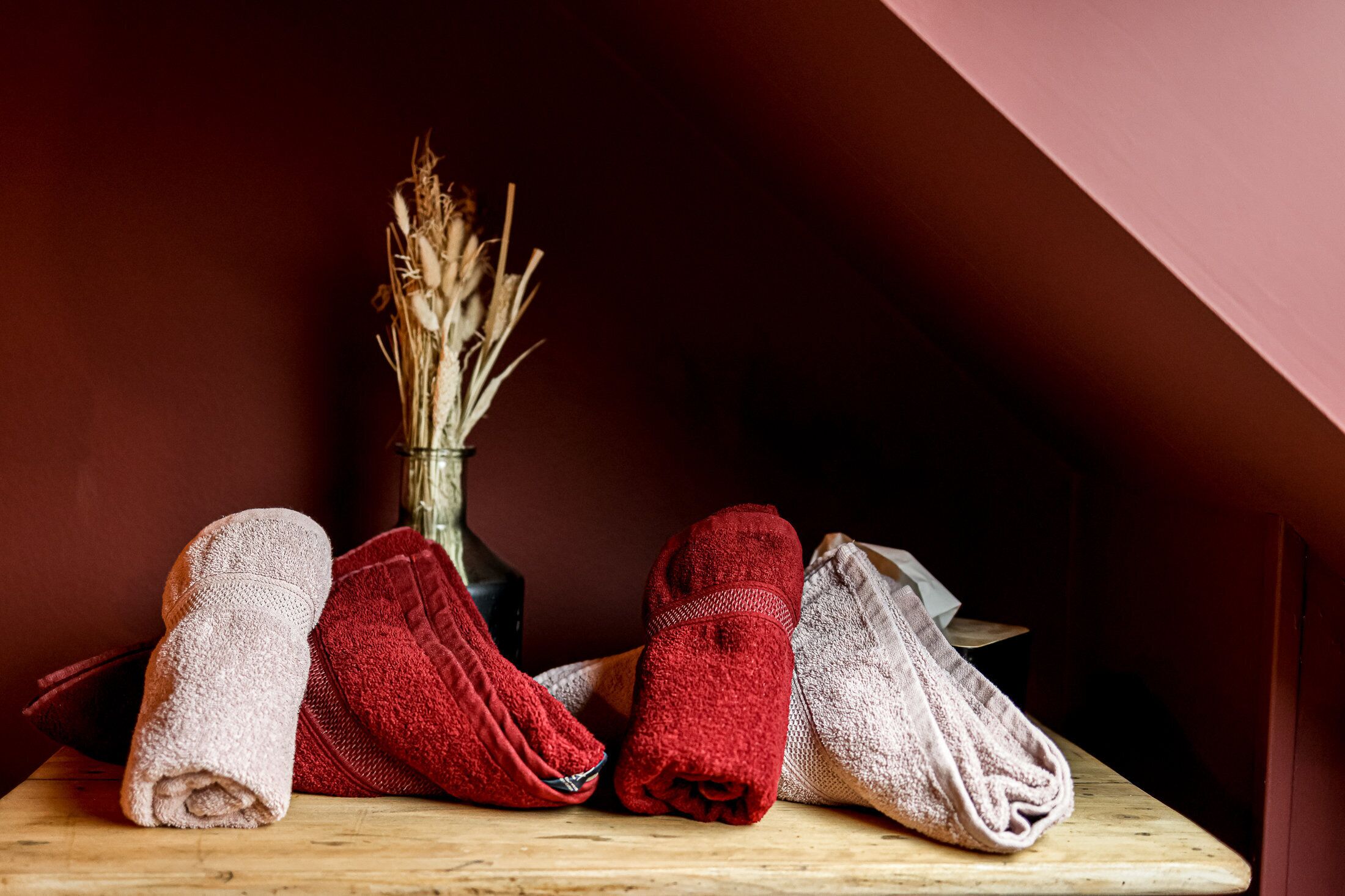 A wooden table with rolled towels in various colors and a vase with dried plants.