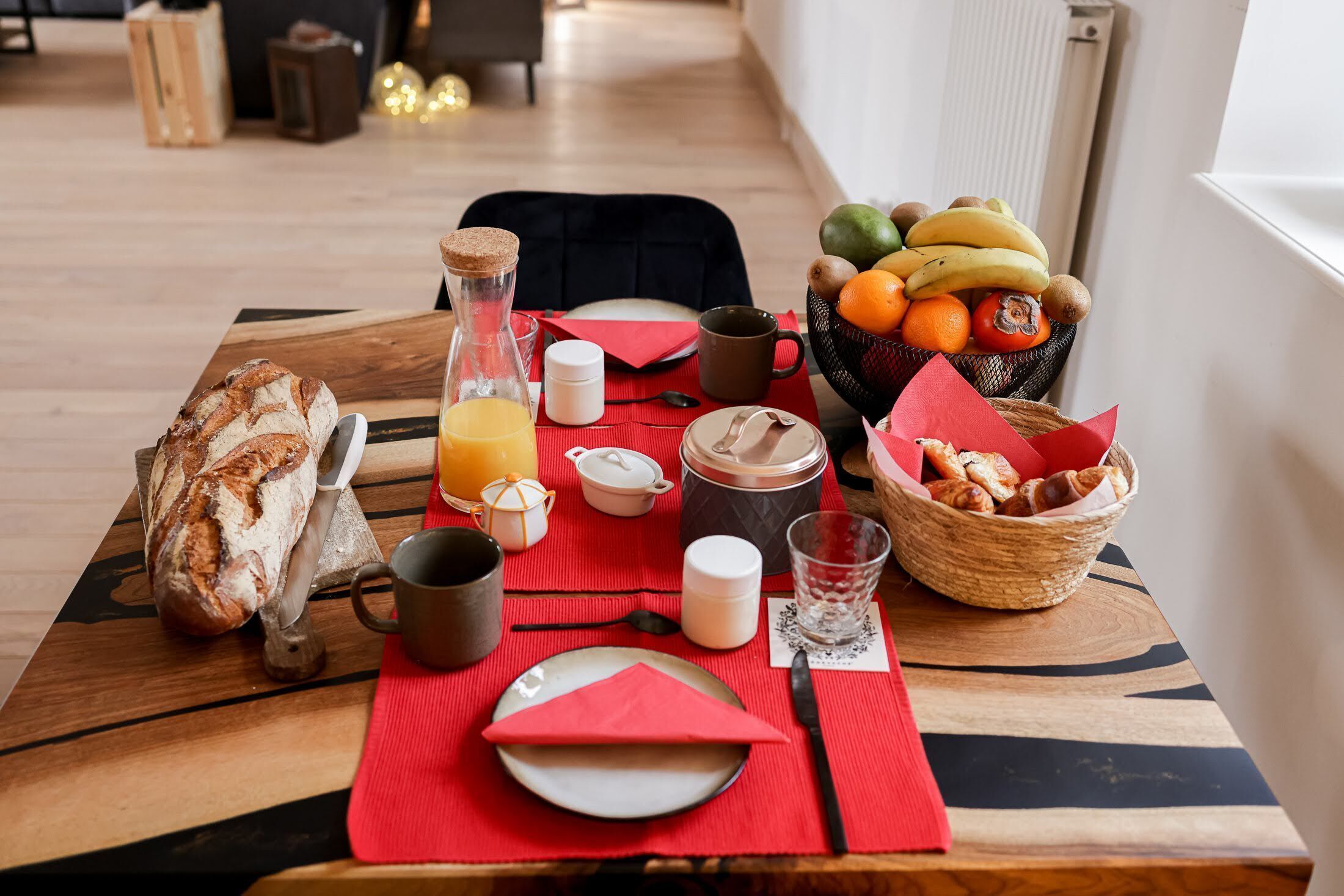 A breakfast setup with bread, fruit, and beverages on a wooden table with a red placemat.