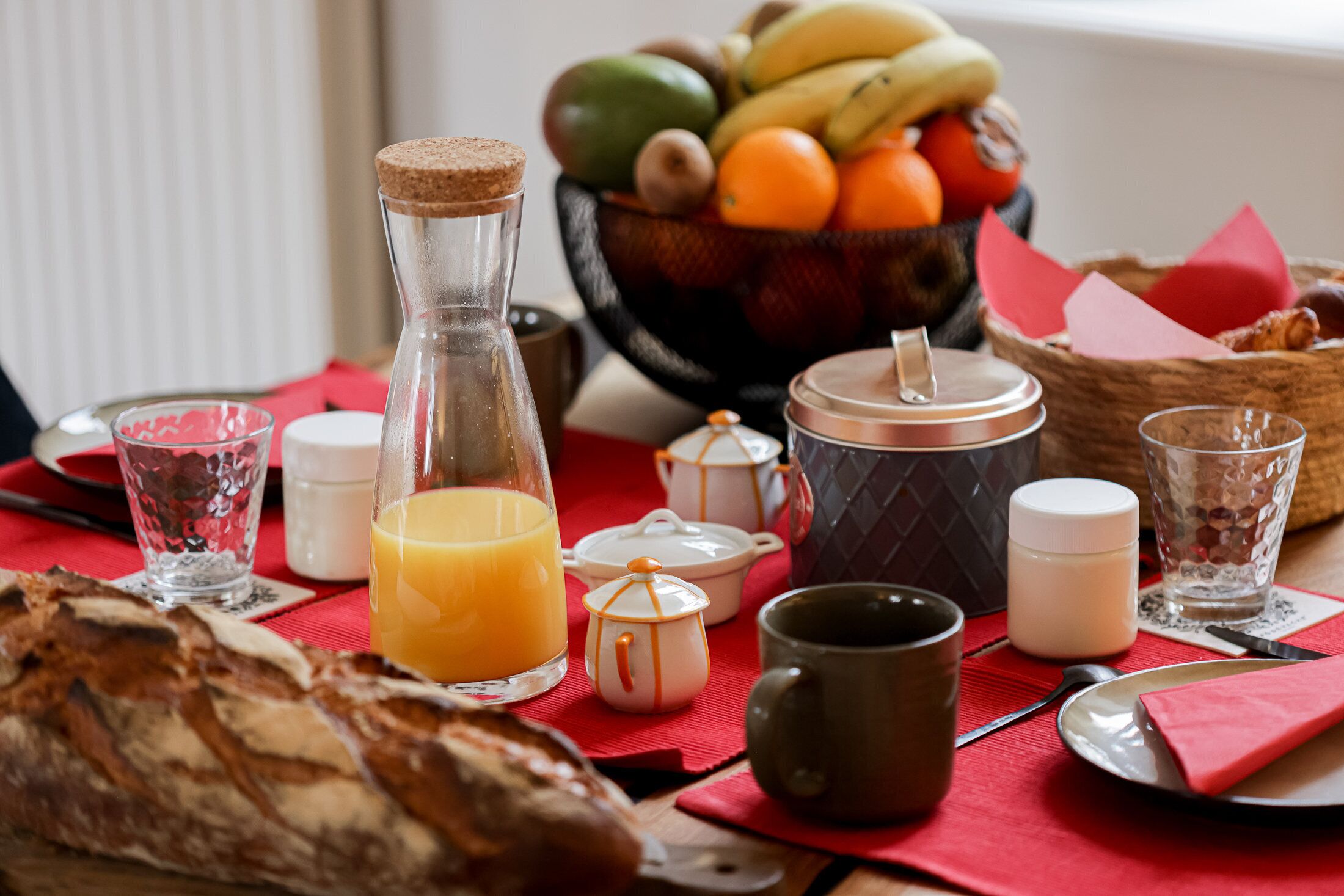 A breakfast setting with bread, a fruit bowl, a bottle of orange juice, and various breakfast items on a red tablecloth.